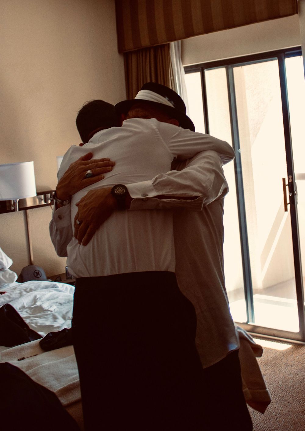 Two men in white dress shirts share an emotional hug in a hotel room during wedding preparations.