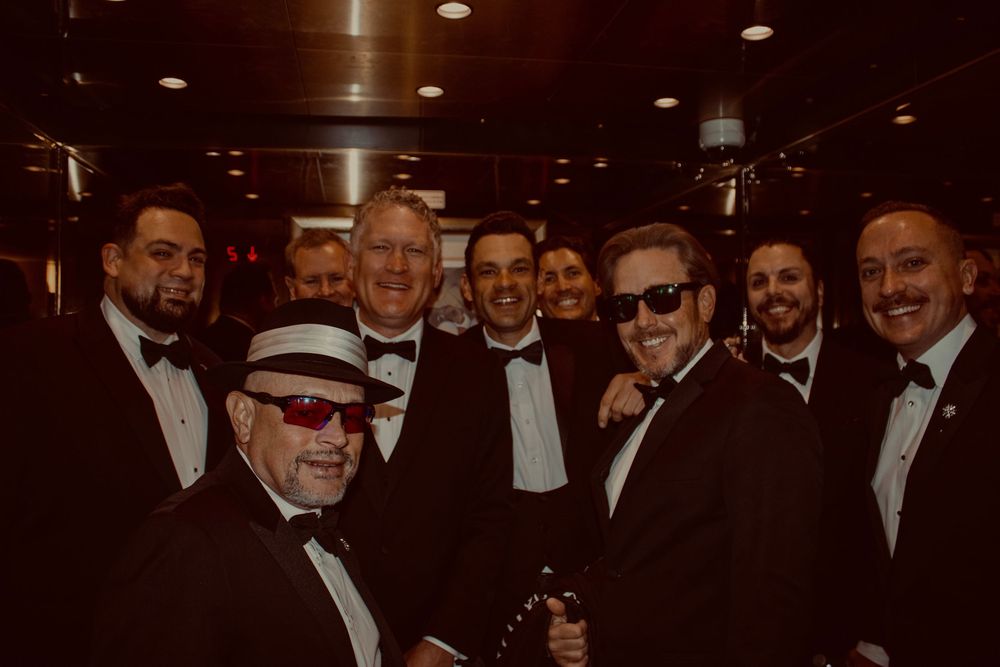 Group of men in black tuxedos and bow ties smiling together at a formal wedding reception.