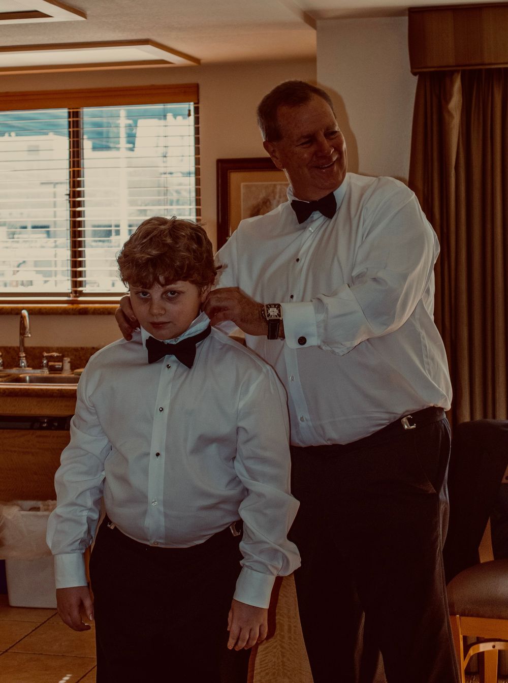 Adult man in tuxedo helping a young boy adjust his bow tie collar while both wear formal black and white attire.