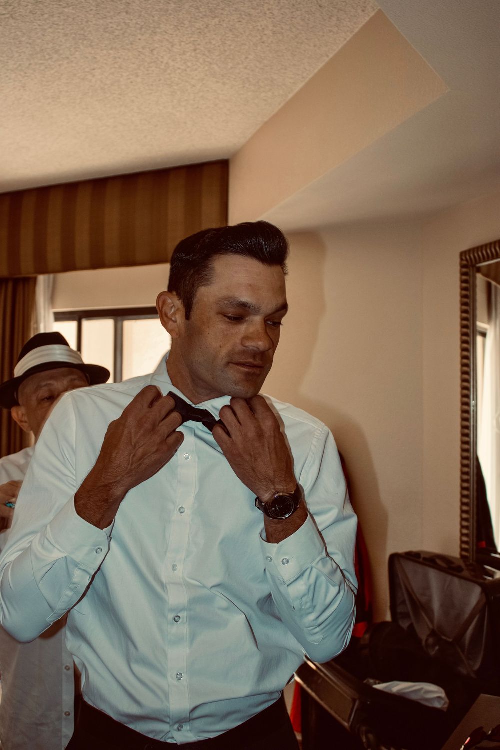 Man in light blue dress shirt adjusting his tie while getting ready in a hotel room.