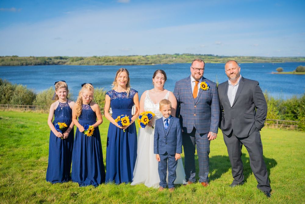 Wedding party in navy blue and white attire poses on grassy hill overlooking scenic lake on sunny day.
