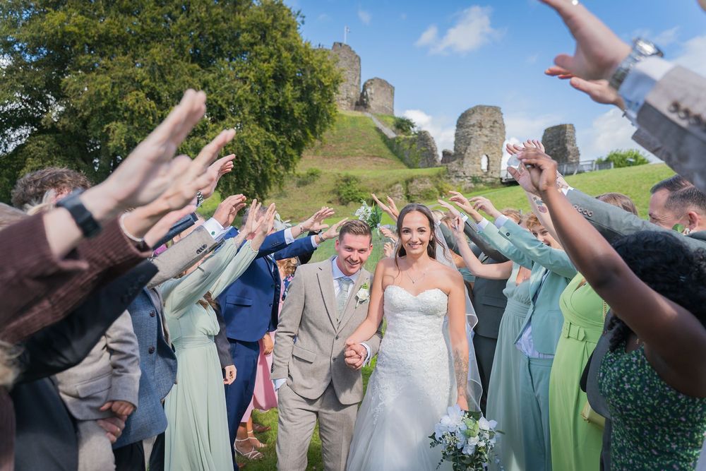 Wedding guests toss confetti and celebrate as newlyweds walk through a joyful receiving line on a sunny day.