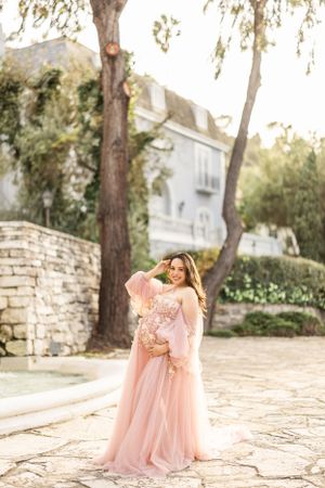 A person in a flowing pink gown stands on stone pavement near trees and a white building in soft sunlight.