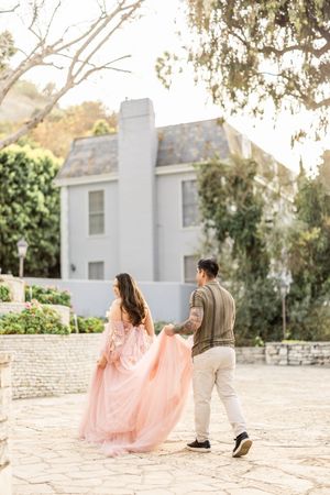Couple walking near white house with flowing pink dress in sunlight.