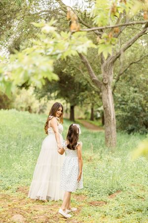 Mother and daughter walking through green meadow in flowing white dresses.