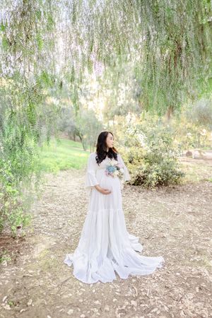 Elegant maternity portrait with flowing white dress under willow tree.