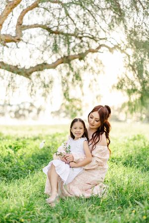 Two people share a tender moment while sitting in lush green grass under a weeping willow tree.