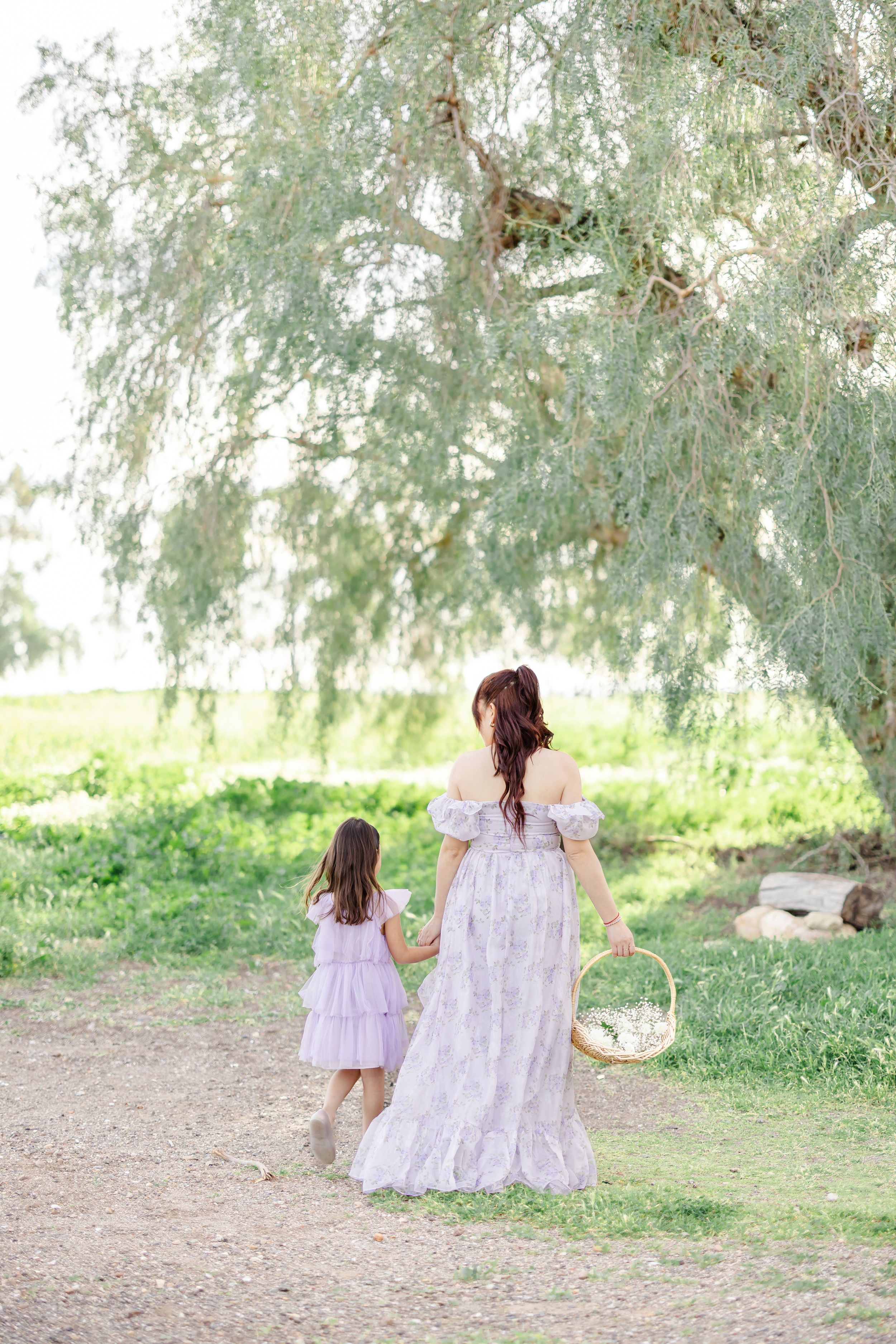 Two figures in matching dresses walk hand in hand along a dirt path under a willow tree.