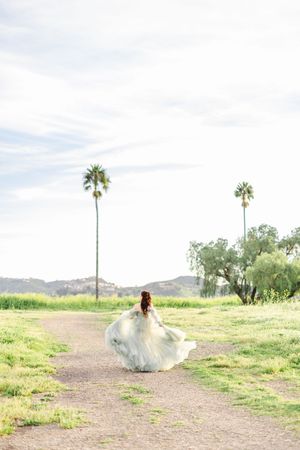 A person in a flowing dress twirls on a path with palm trees and mountains in the background.