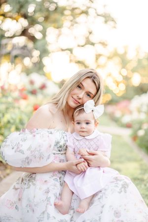 Mother and baby in matching floral dresses pose together in a dreamy garden setting.