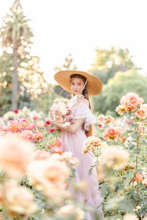 Figure wearing straw hat explores garden of peach and pink roses at sunset.