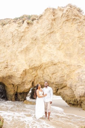 A couple in white outfits stands together on a beach with towering golden cliffs behind them.