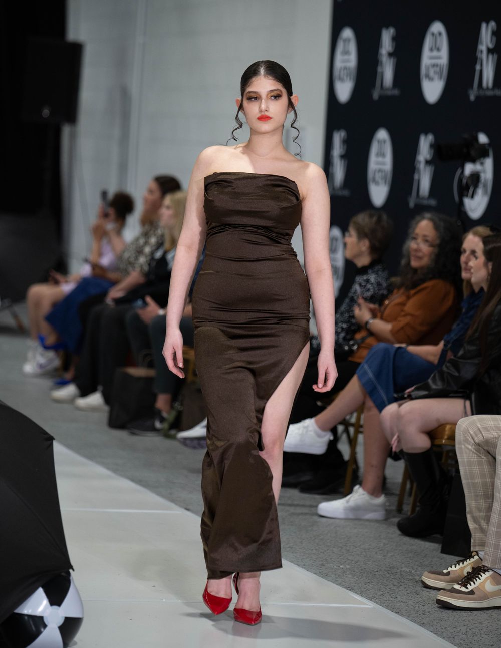 Fashion model in brown strapless dress with side slit walking runway in red heels.