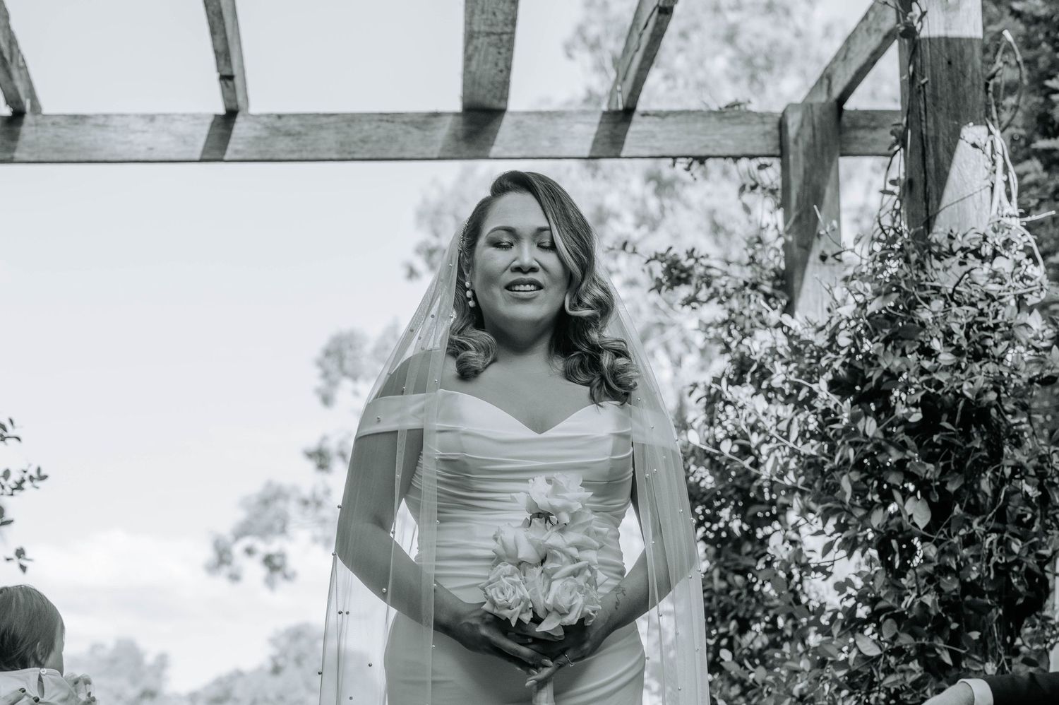 Black and white portrait of a radiant bride holding a bouquet during an outdoor ceremony.
