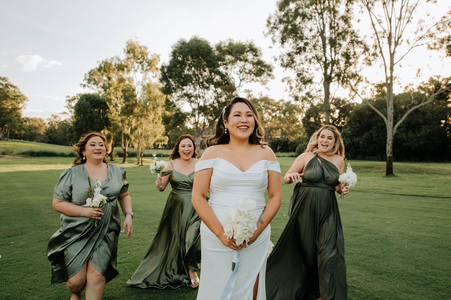 Bridesmaids in sage green dresses running joyfully behind bride on grassy lawn.