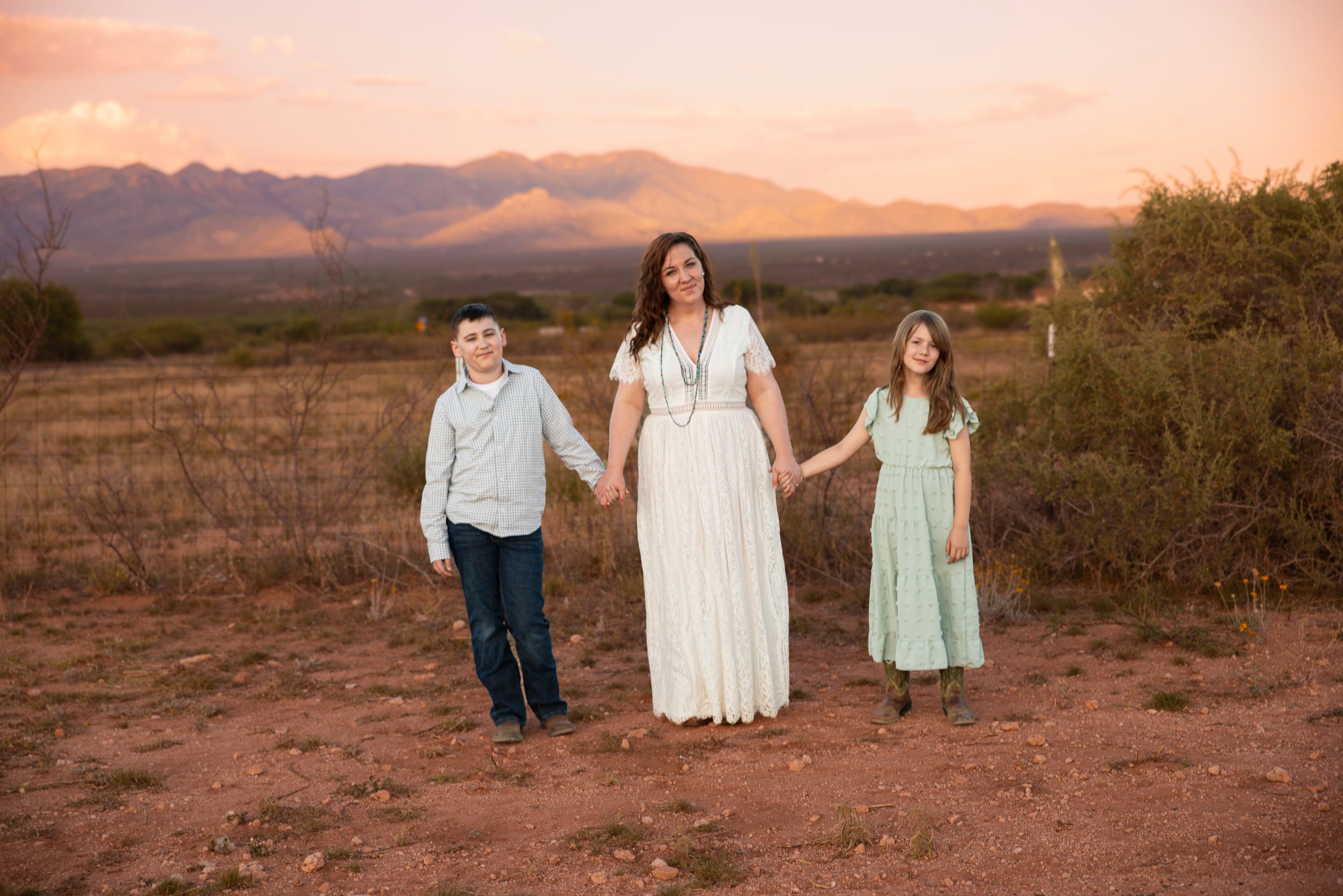 Mother in a long white dress holds hands with a boy and a girl in a desert field at sunset with mountains in the background.