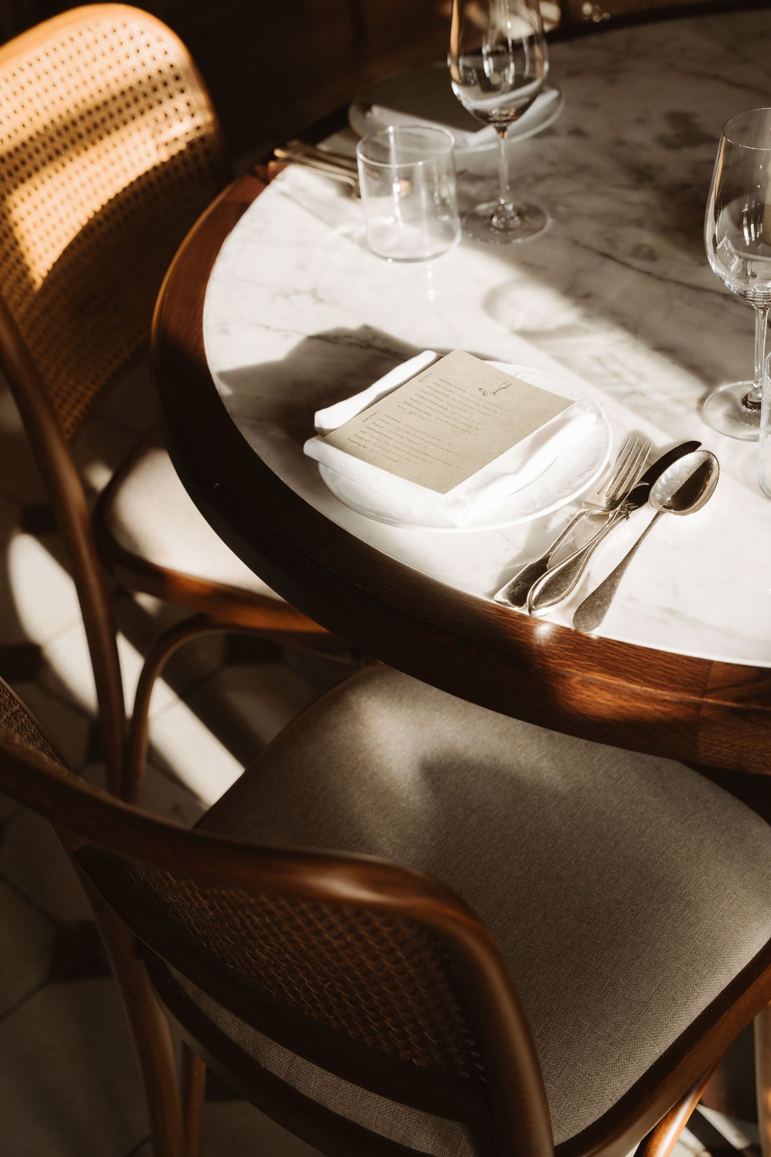 Elegant restaurant table setting with wooden bistro chairs, white tablecloth, and wine glasses in moody lighting.
