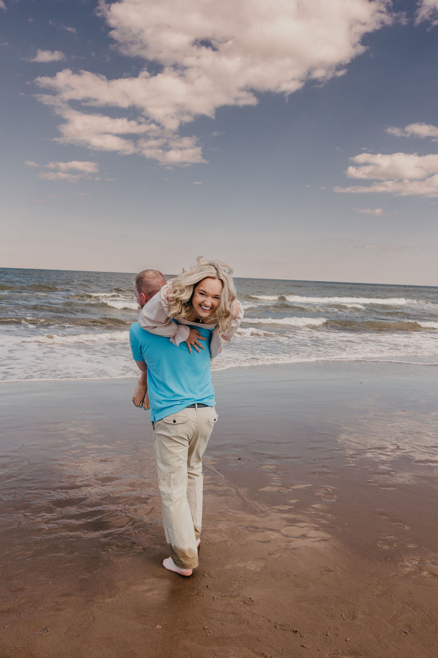 Person in a blue shirt and khaki pants dances joyfully on a beach at sunset.