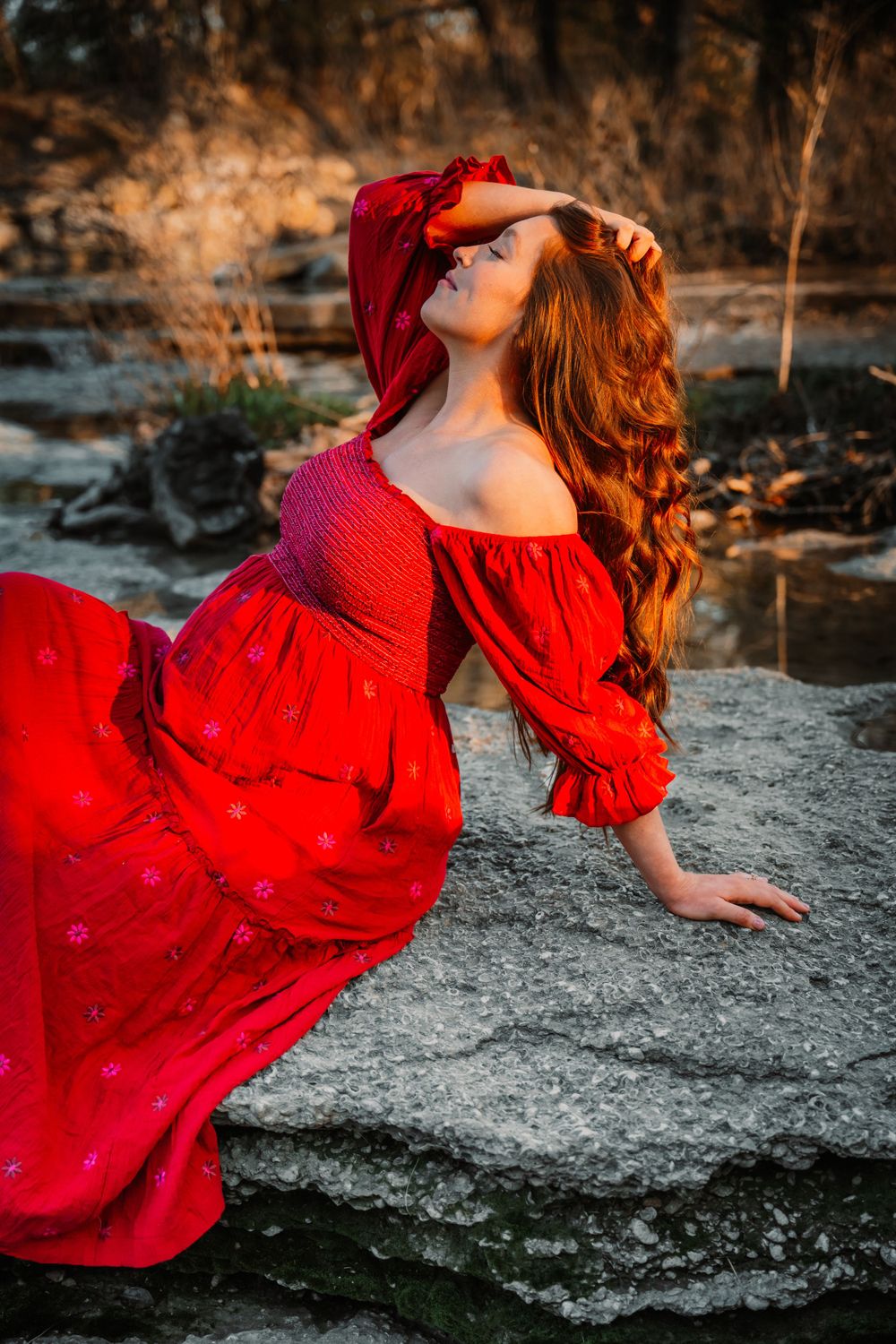 Pregnant woman in red dress posing on rocks at a waterfall during outdoor maternity photo session in Dallas Fort Worth