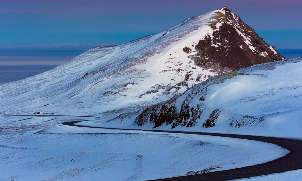 Snow-covered mountain peak with winding road against deep blue twilight sky.