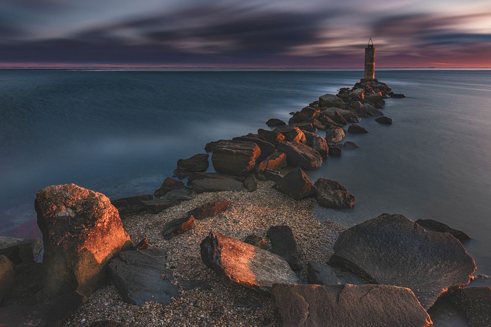 Rocky jetty extends into calm ocean waters during dramatic sunset with warm lighting.