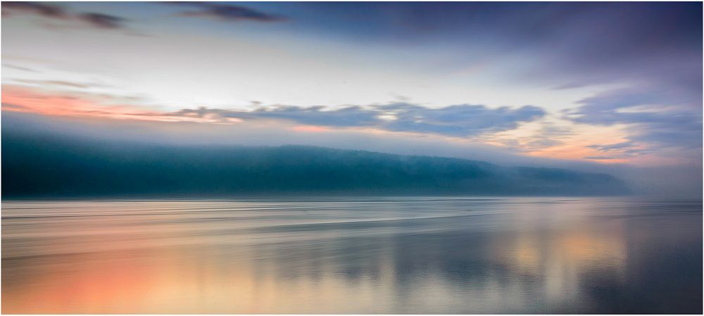 Serene seascape with pastel sunrise colors reflecting on calm waters under cloudy sky.