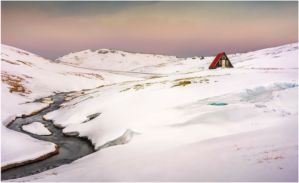 Small red cabin beside winding stream in vast snow-covered winter landscape.