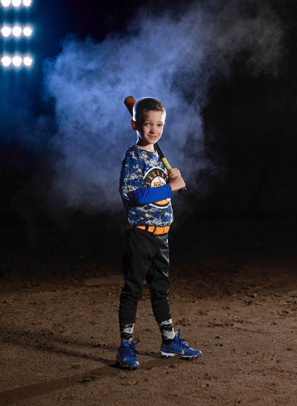 Young athlete poses confidently in blue sports attire against dramatic smoky backdrop with stadium lighting.