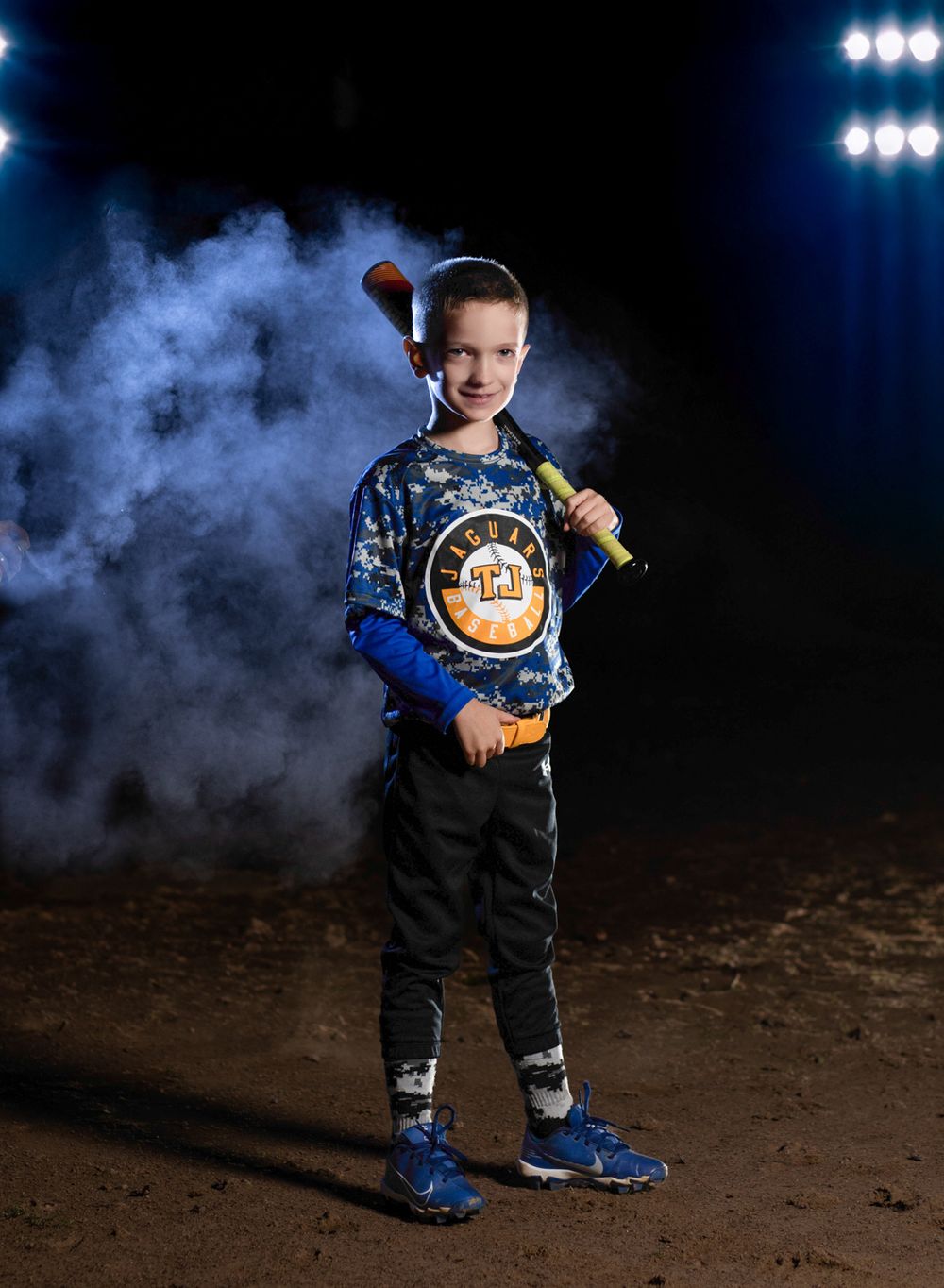 Youth baseball player poses with bat in team uniform against dark background with atmospheric smoke and stadium lights.