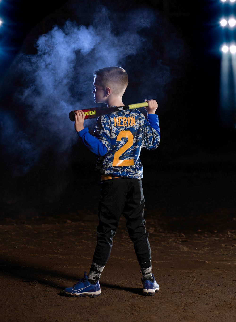 Young baseball player in blue jersey number 2 poses with bat against dark background with stadium lights and smoke.
