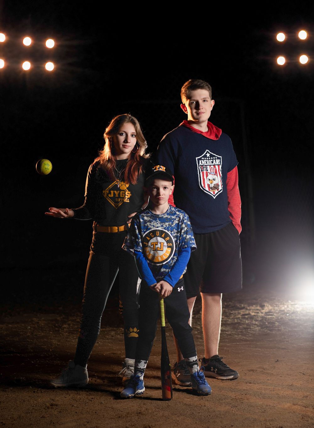 Baseball players stand together at night under bright lights on a dirt field in athletic apparel.