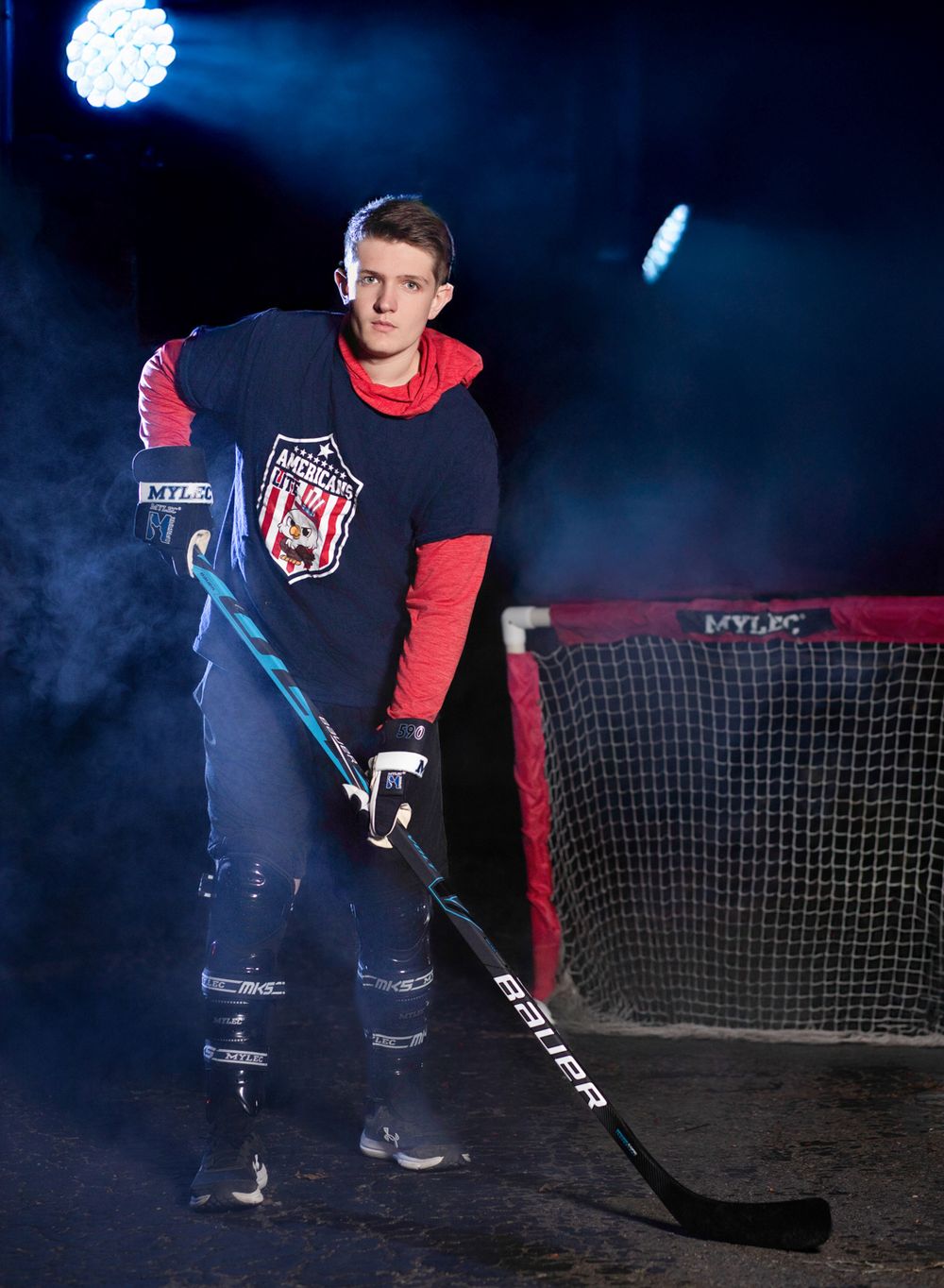 Hockey player poses in dark blue and red uniform with stick on ice rink with dramatic lighting and fog effects.