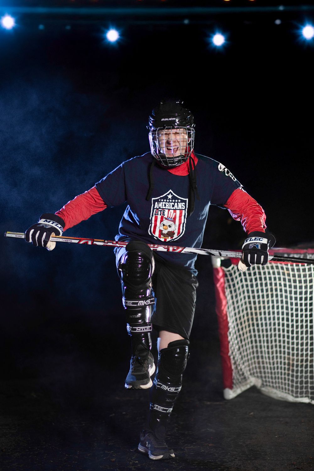 Hockey player in black and red uniform with USA-themed logo stands by goal net under dramatic blue lighting.