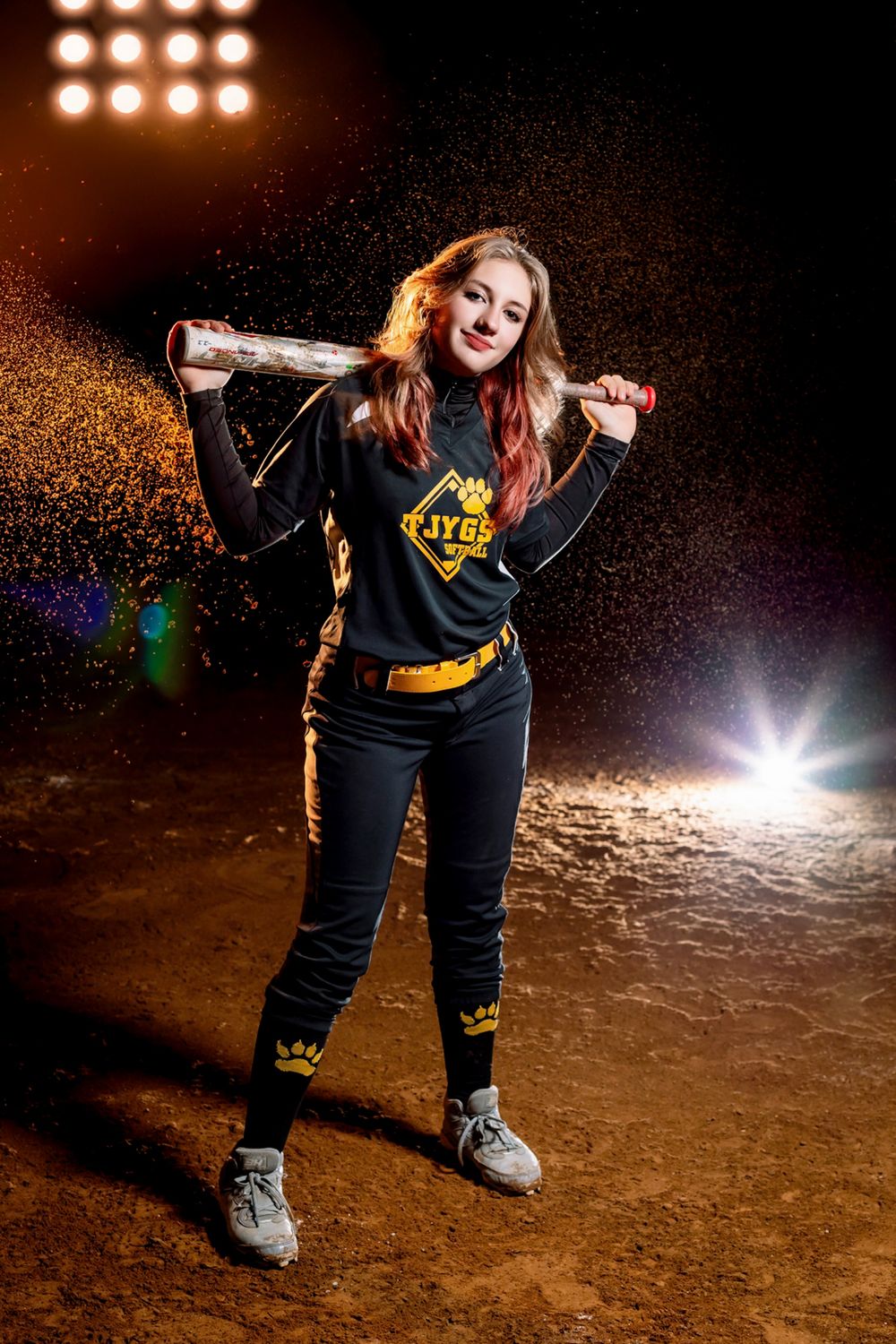 A player in a softball uniform poses with a bat on a dirt field at night while golden glitter sparkles in the air.