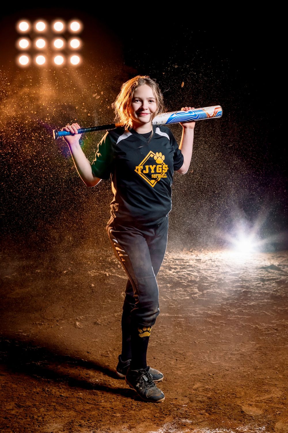 Softball player poses with bat across shoulders on dirt field at night under stadium lights with dramatic dust effect.