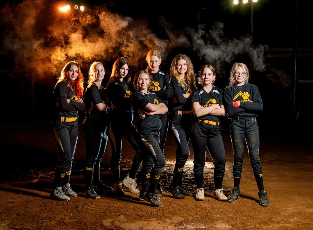 Dramatic softball team photo at night with smoke effects and matching dark uniforms on a dirt field.