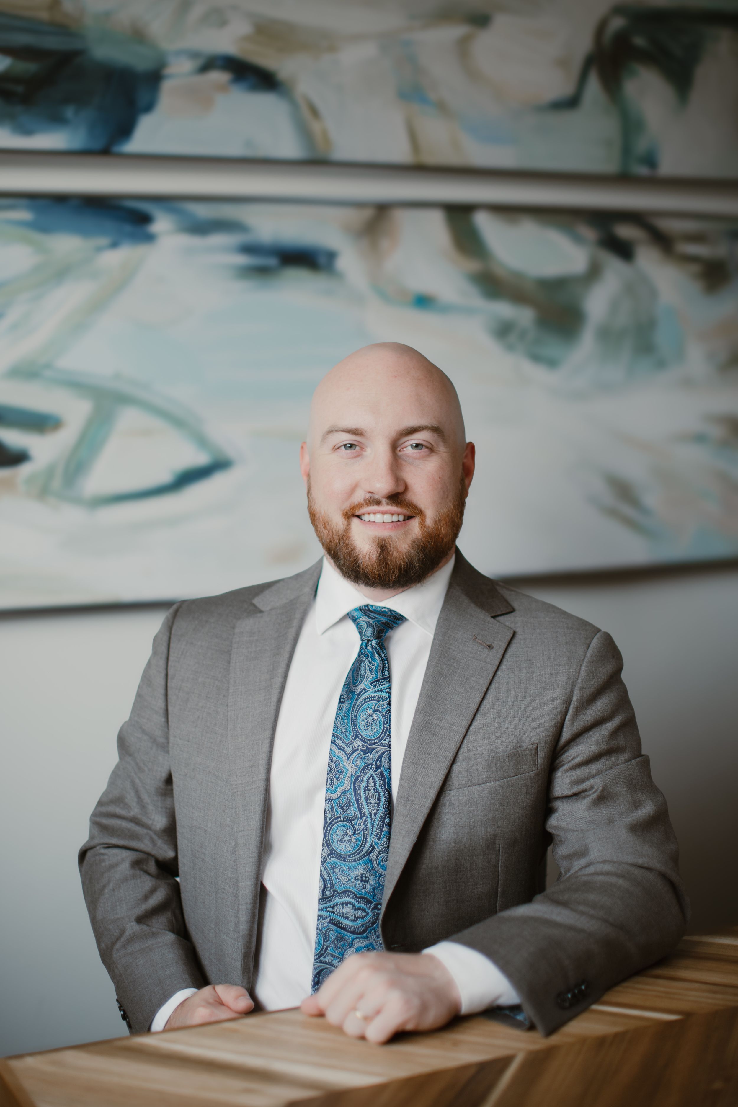 Professional portrait of person in gray suit and blue paisley tie sitting at wooden desk.