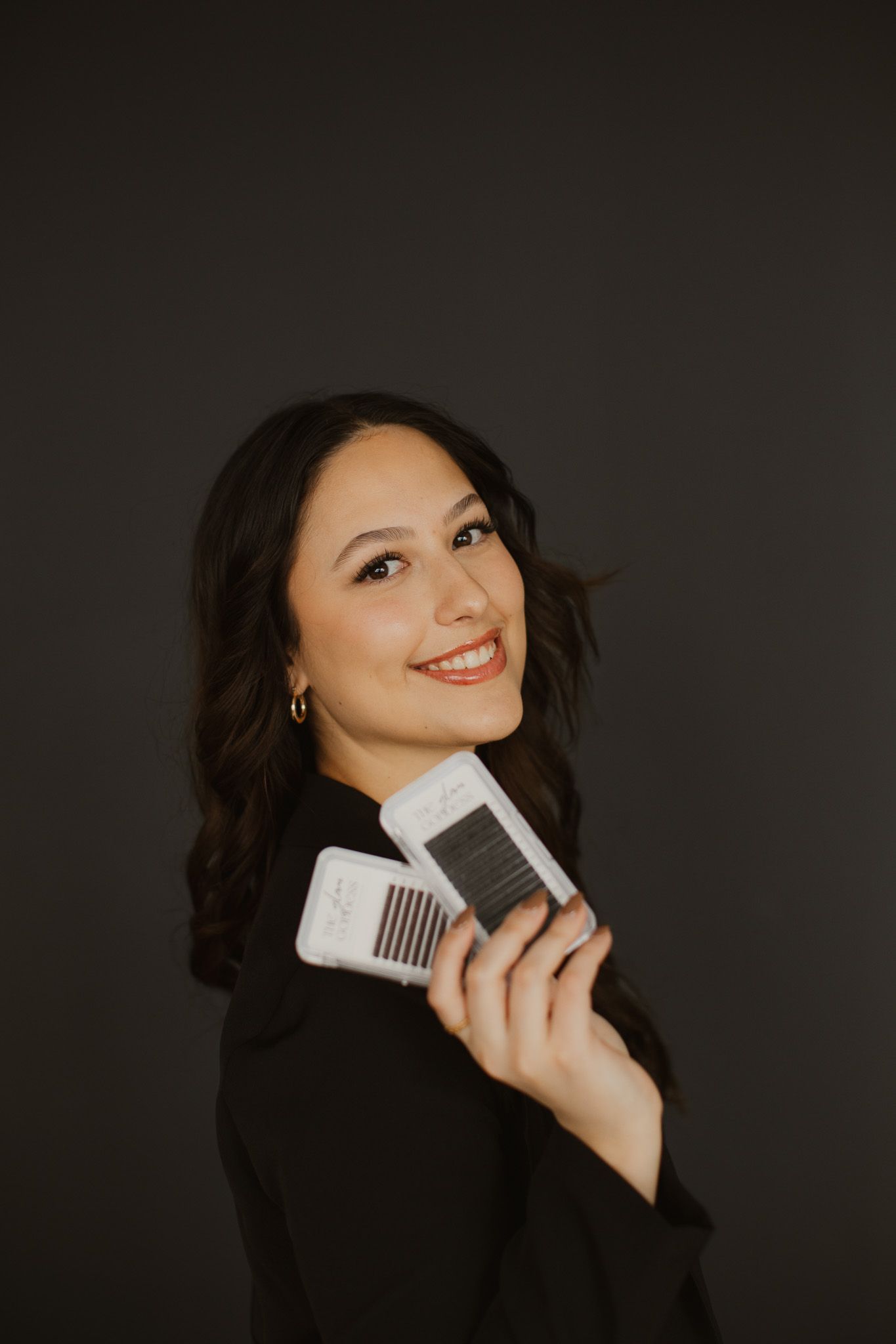 Professional beauty artist holding eyelash extension fans while smiling against dark background.