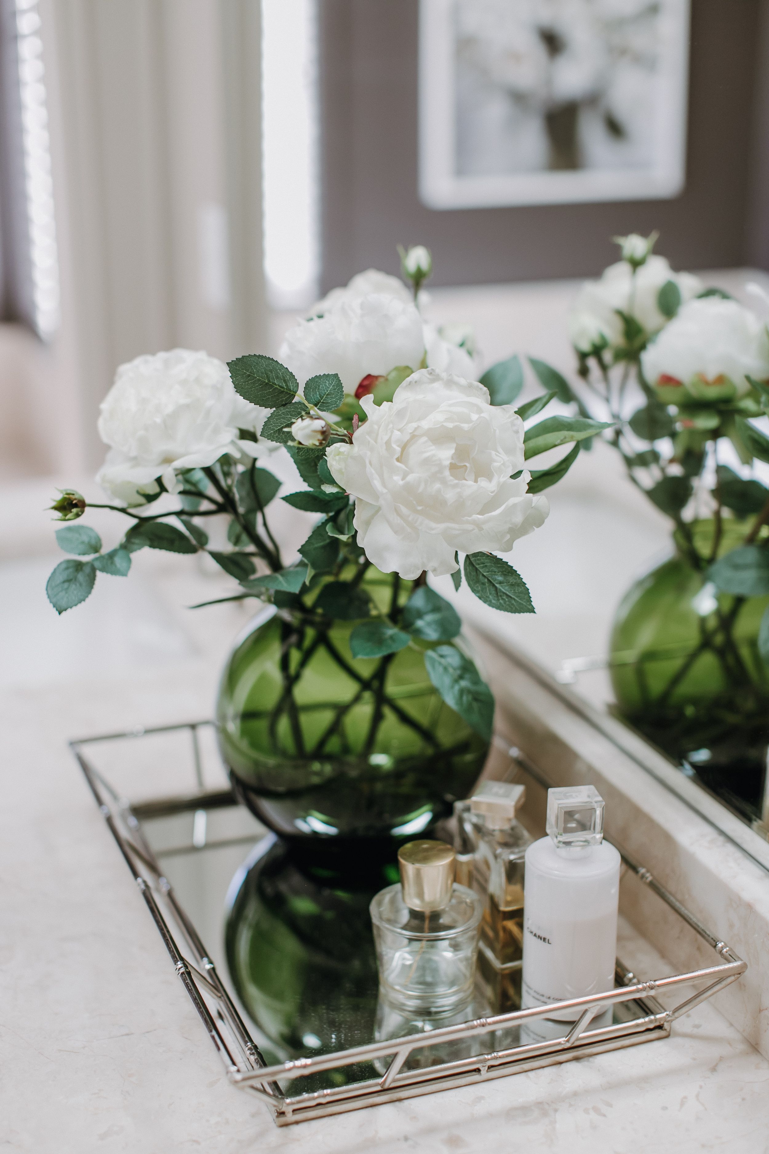 Elegant white roses in green glass vase with decorative tray and bottles on marble surface.
