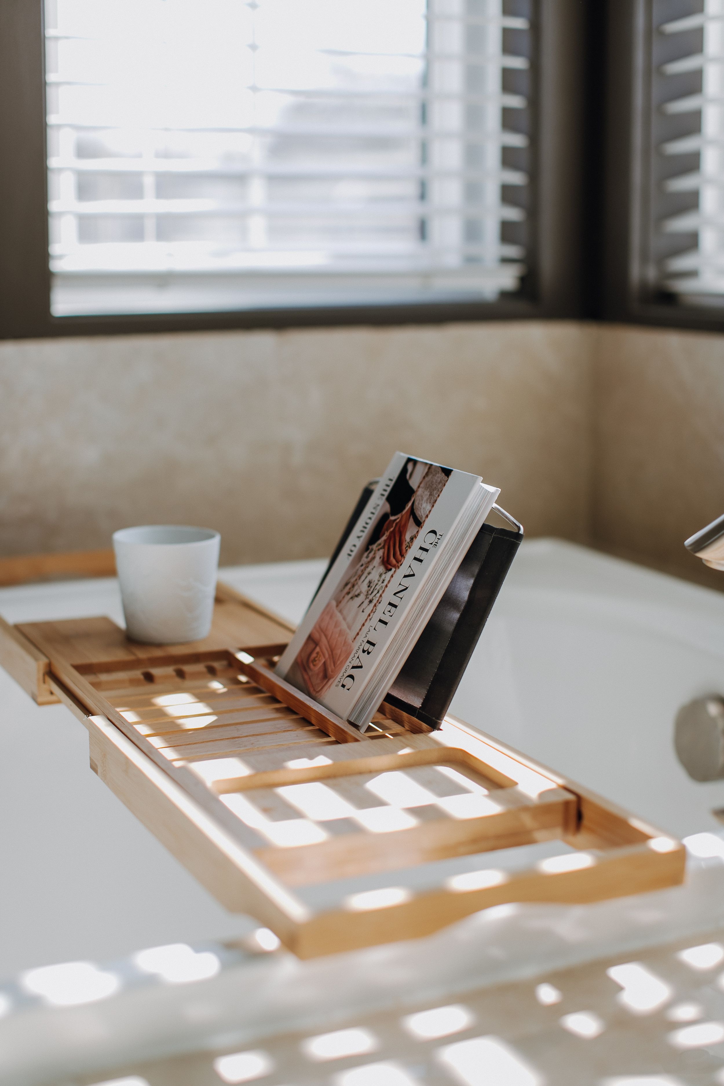 Wooden bathtub caddy with a book and glass catches morning light through window blinds.