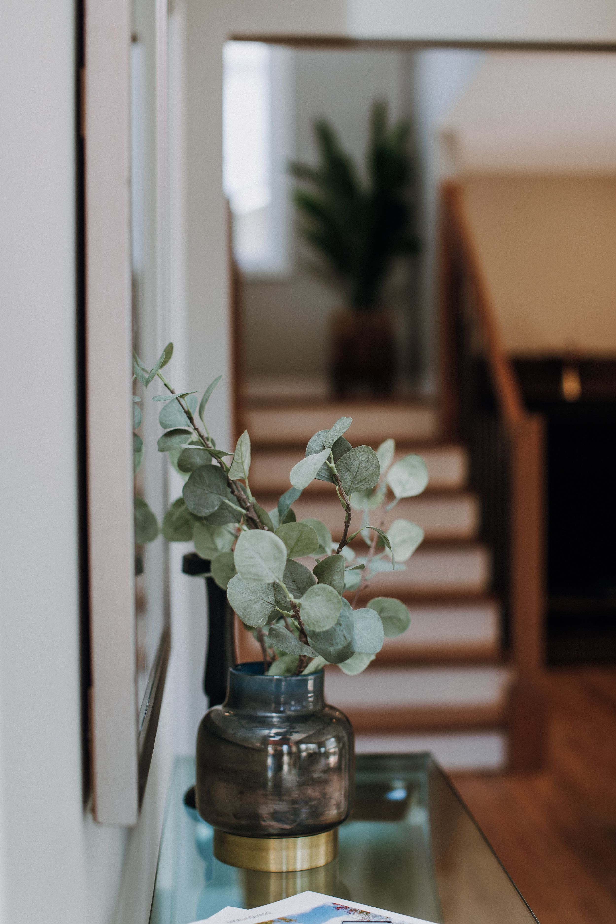 Eucalyptus branches in a smoky glass vase with brass accent sit on a glass shelf near stairs.