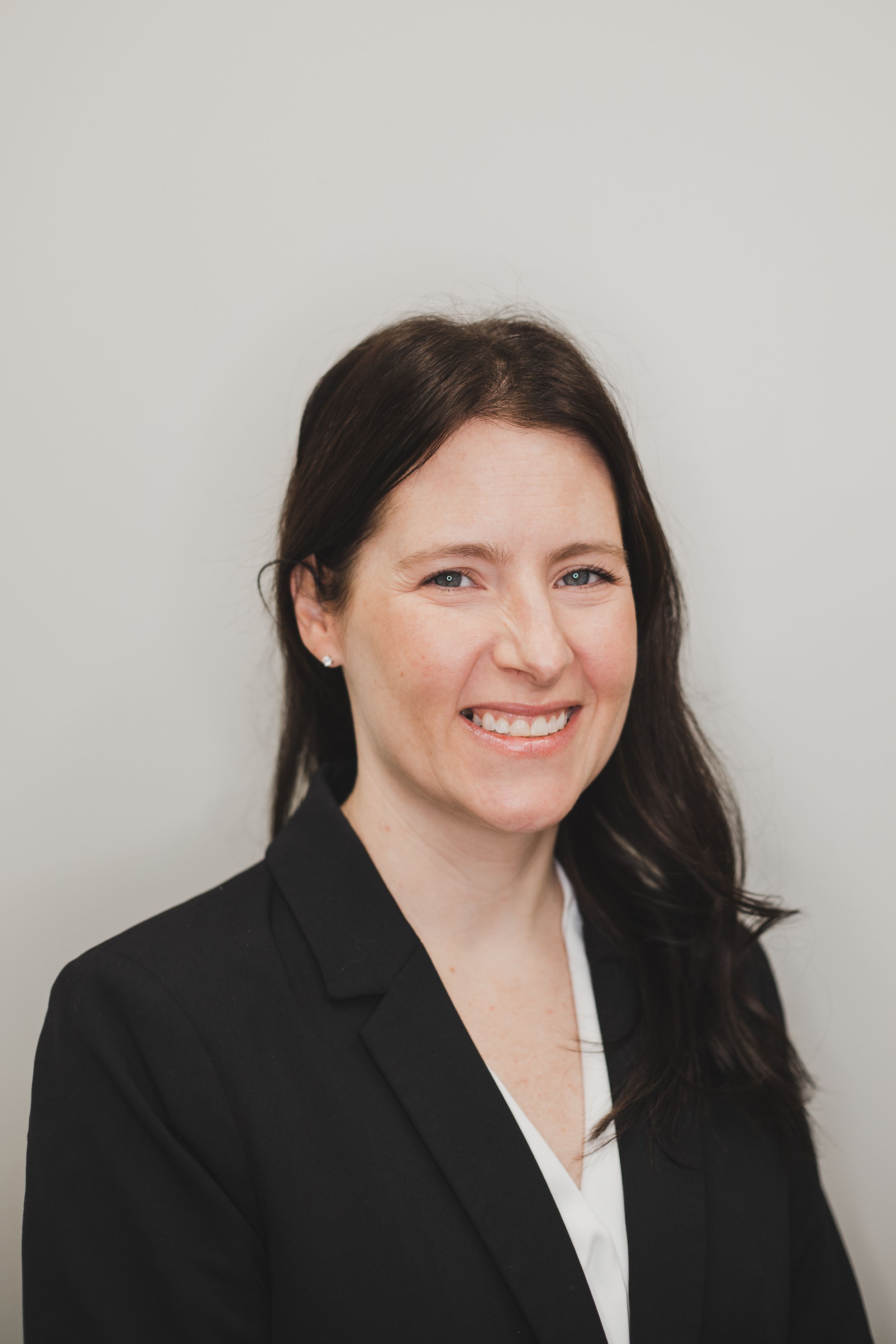 Professional business portrait of a person in a black blazer and white blouse against a gray background.