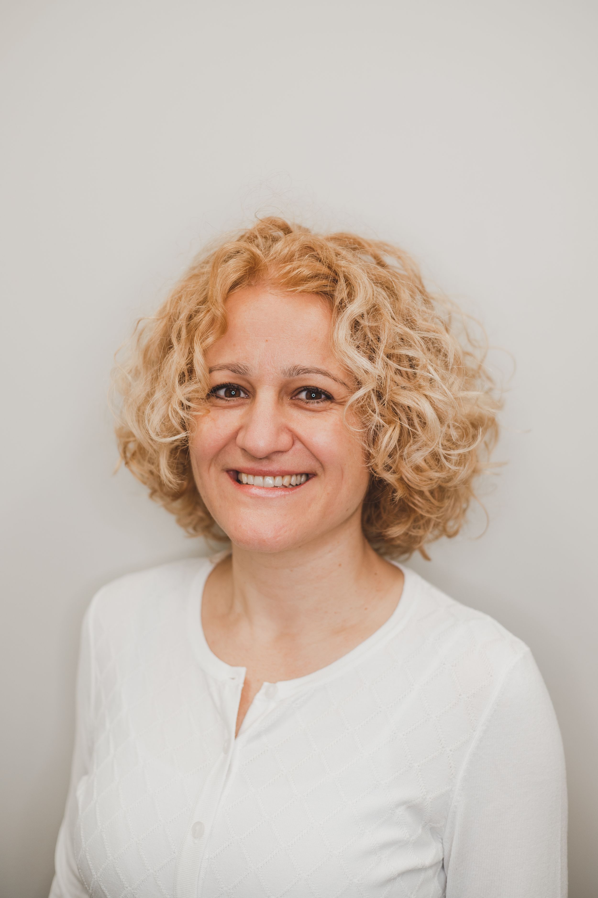 Professional headshot of a person with curly blonde hair wearing a white top against a neutral background.