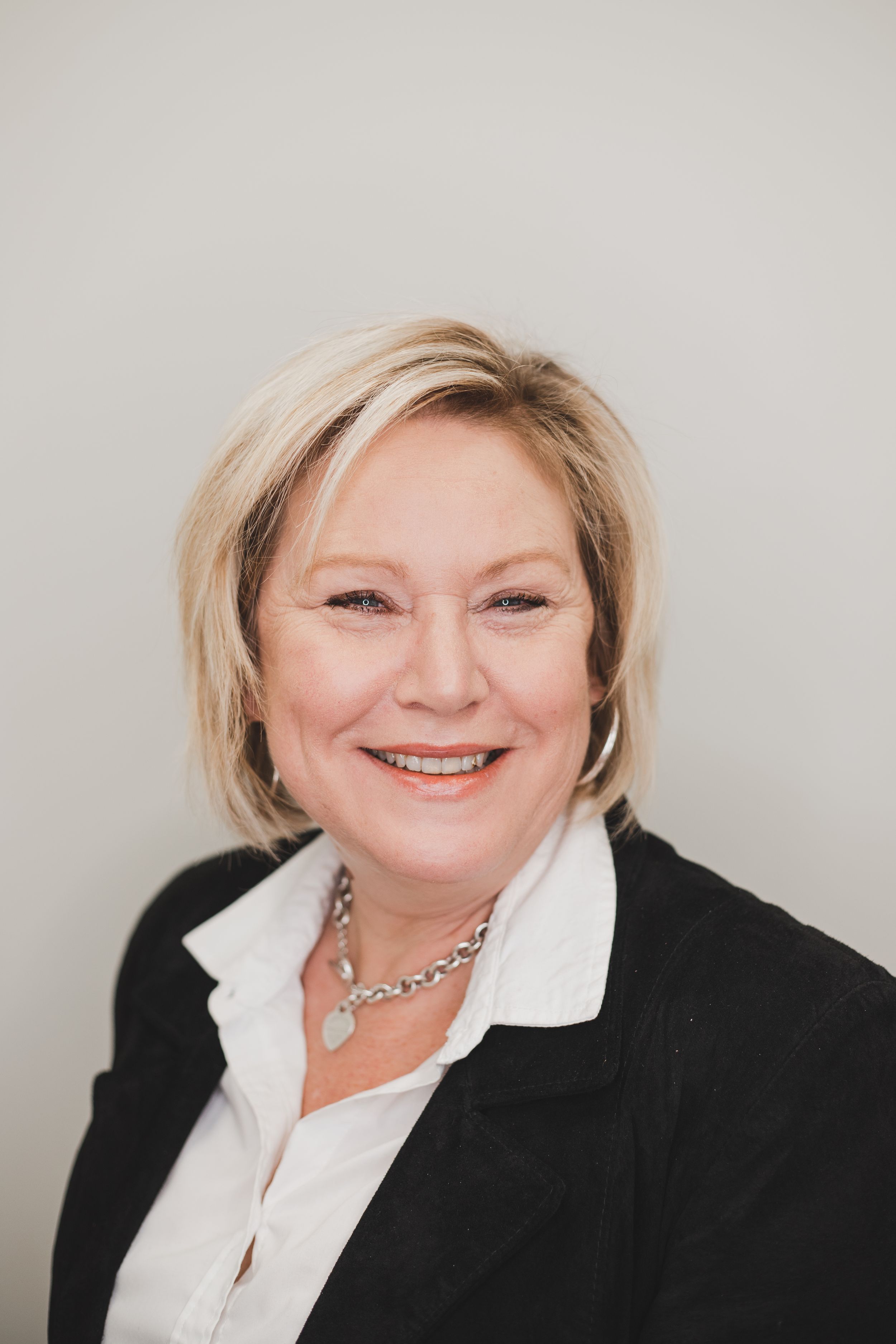 Professional headshot of a person in business attire with a pearl necklace against a neutral backdrop.