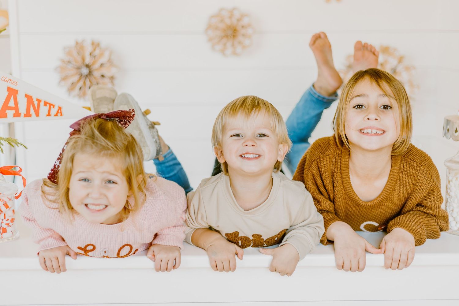 Three smiling children lying on a white surface with festive holiday decorations in the background.