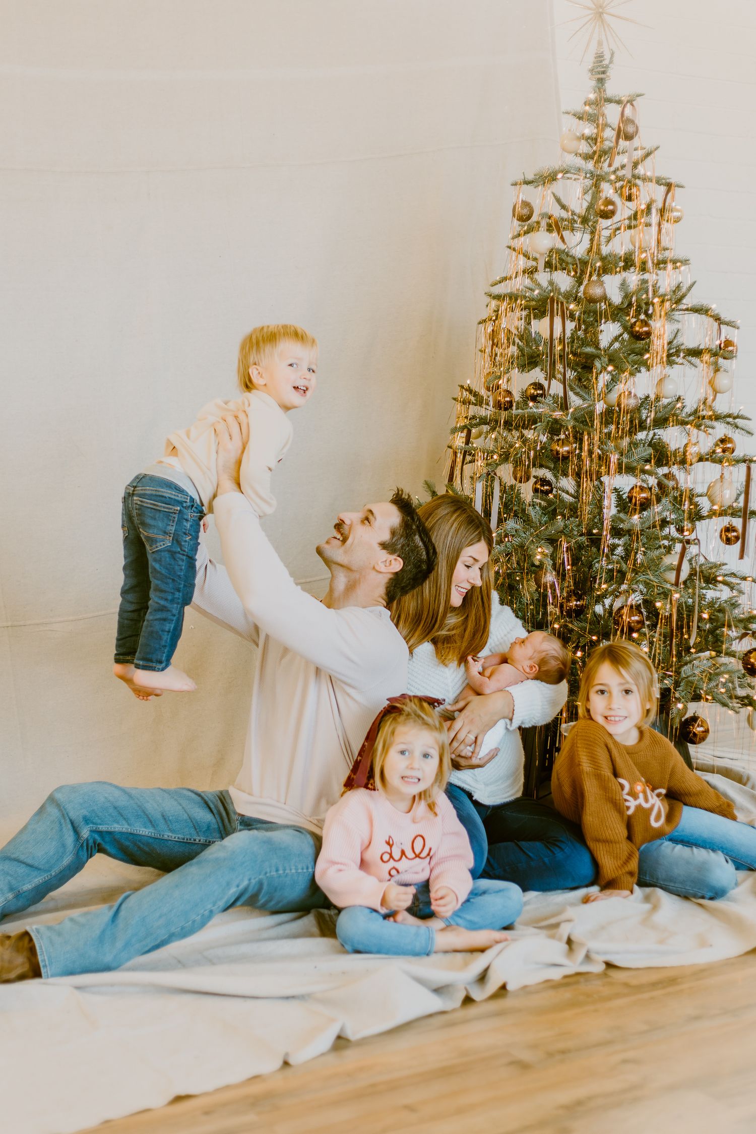 Family of six poses joyfully by decorated Christmas tree, parents lifting toddler while holding newborn, children in festive attire.