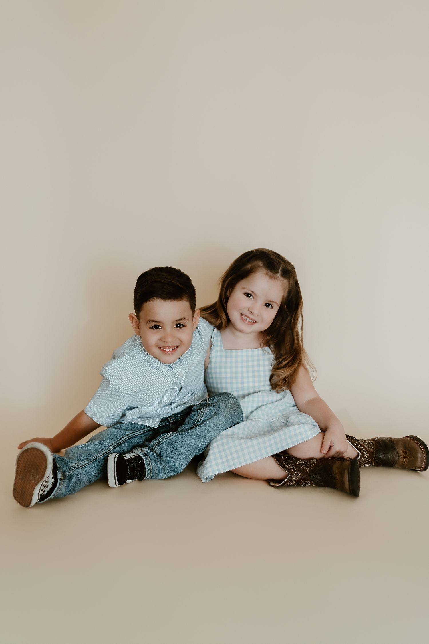 Two young children smiling and posing together on a studio floor, boy in blue shirt and jeans, girl in gingham dress.