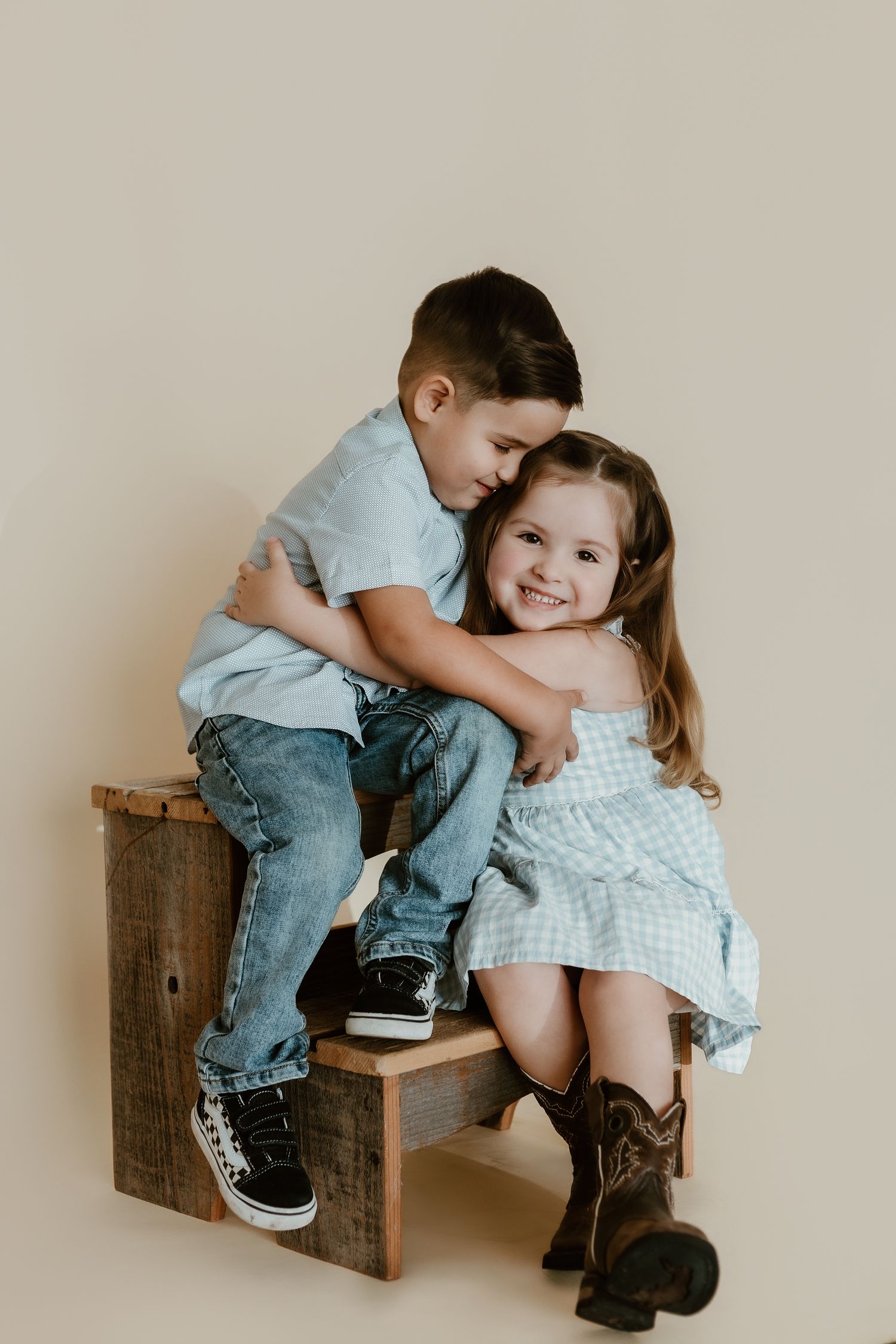 Two young siblings share a sweet hug on a wooden stool, girl in blue dress and cowboy boots, boy in jeans.