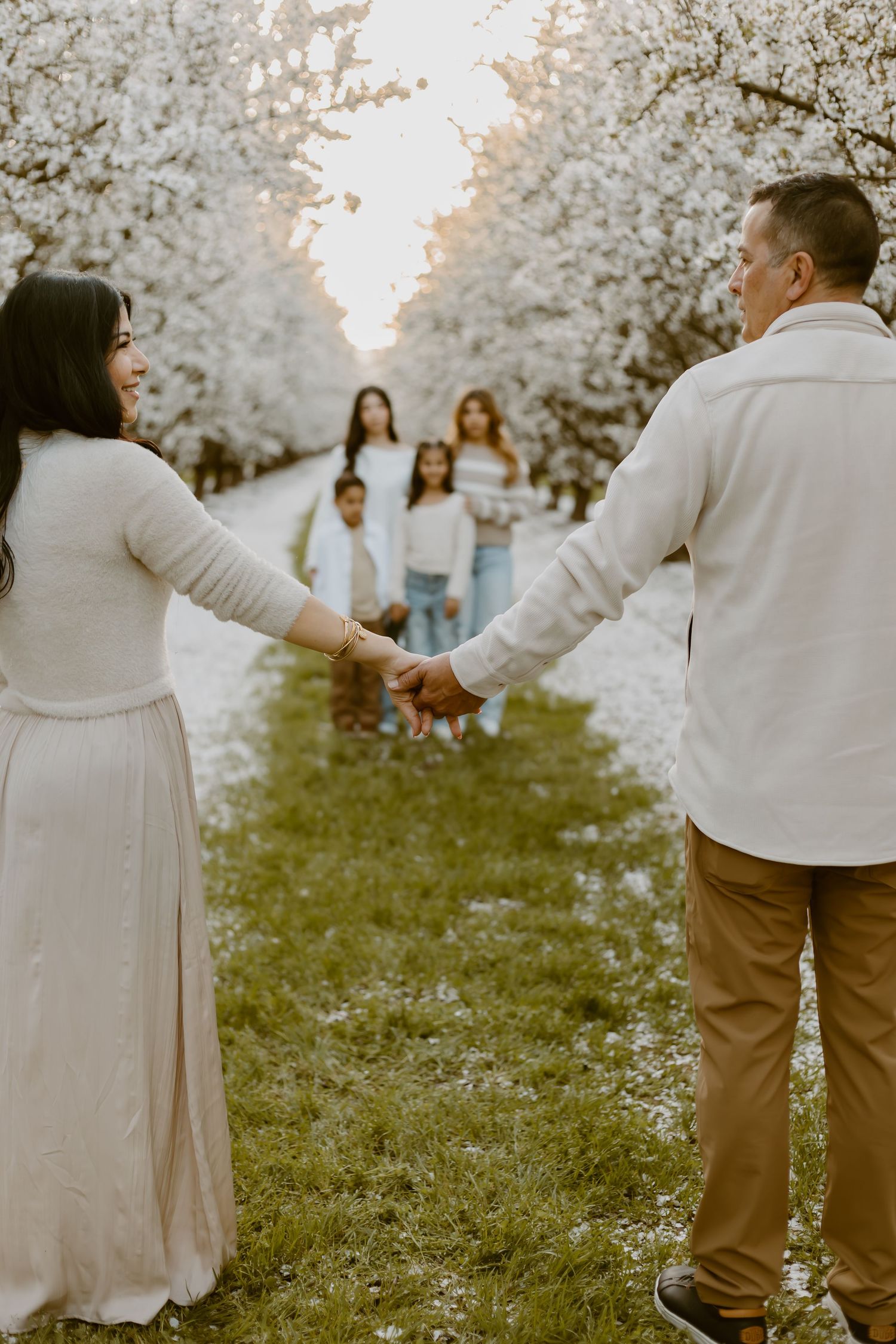 A family holds hands along a blooming cherry blossom tree path, with children walking toward their parents in soft natural light.