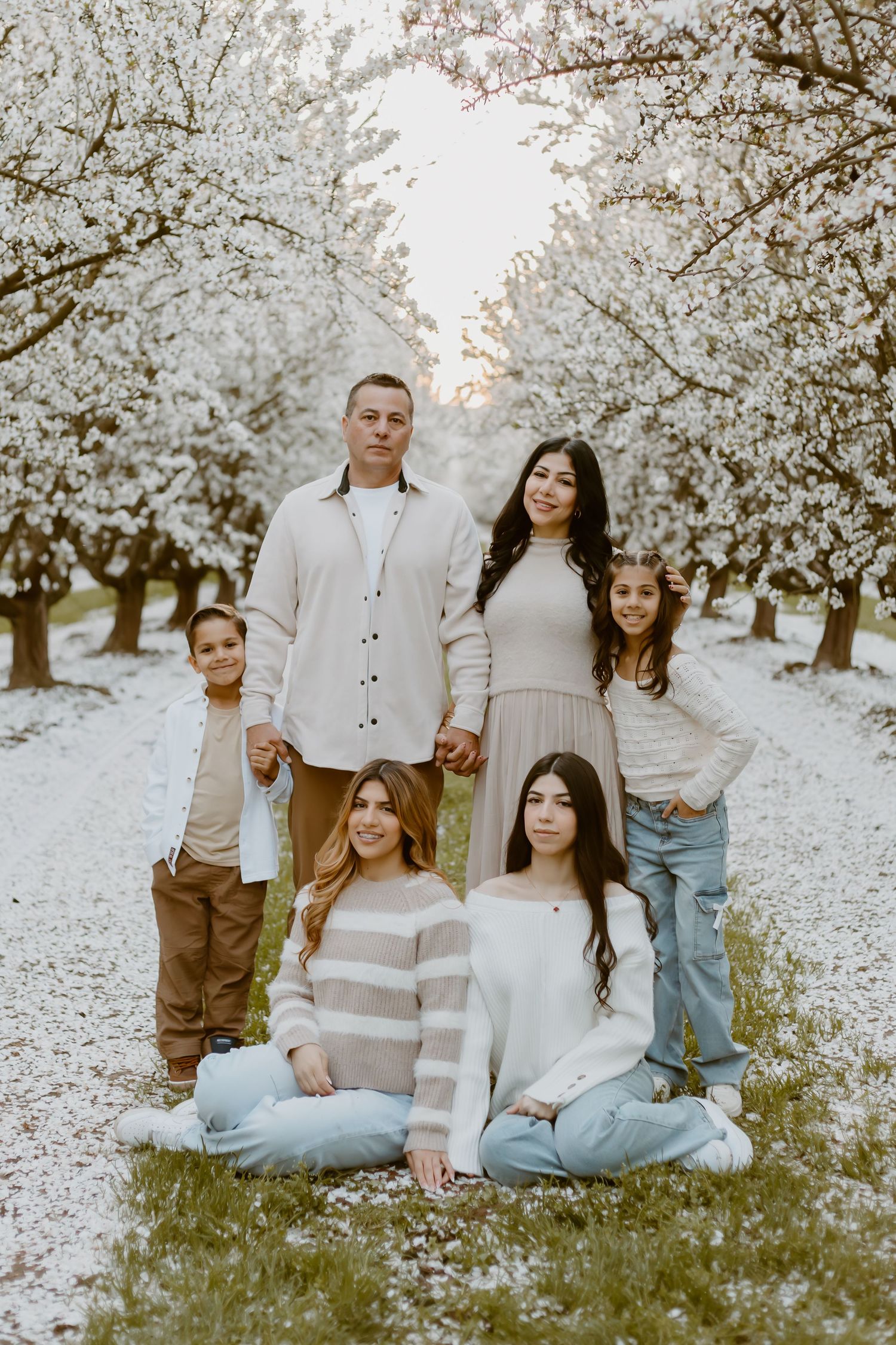 Family of five posing among blooming white cherry blossom trees, wearing neutral tones in a serene orchard setting.